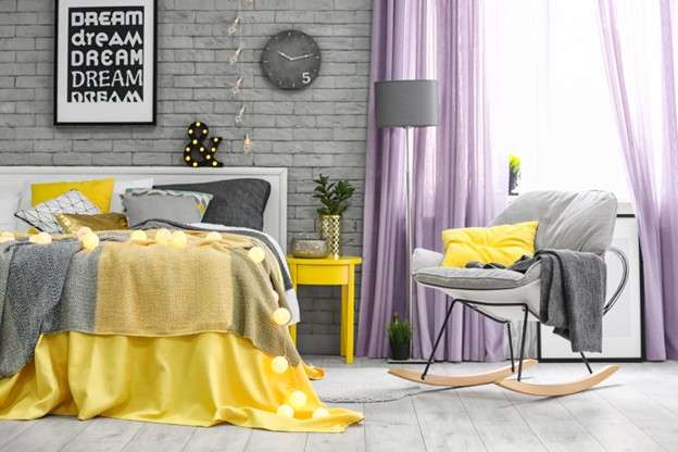A bedroom with a bed and rocking chair draped in yellow, grey, and purple decor