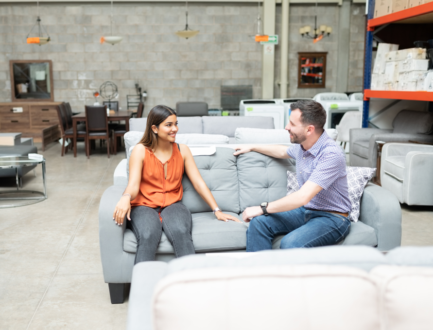 A person sits on a grey sofa at a furniture store while speaking to a sales rep