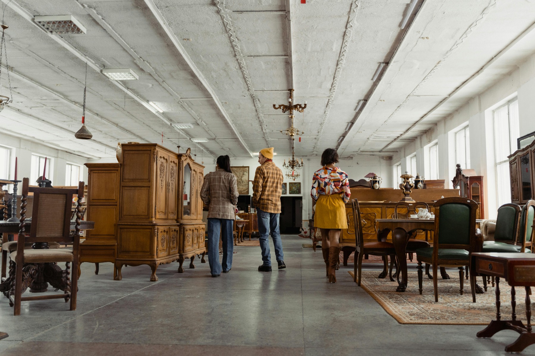 Three people walking through a spacious antique furniture showroom filled with wooden cabinets, tables, and chairs