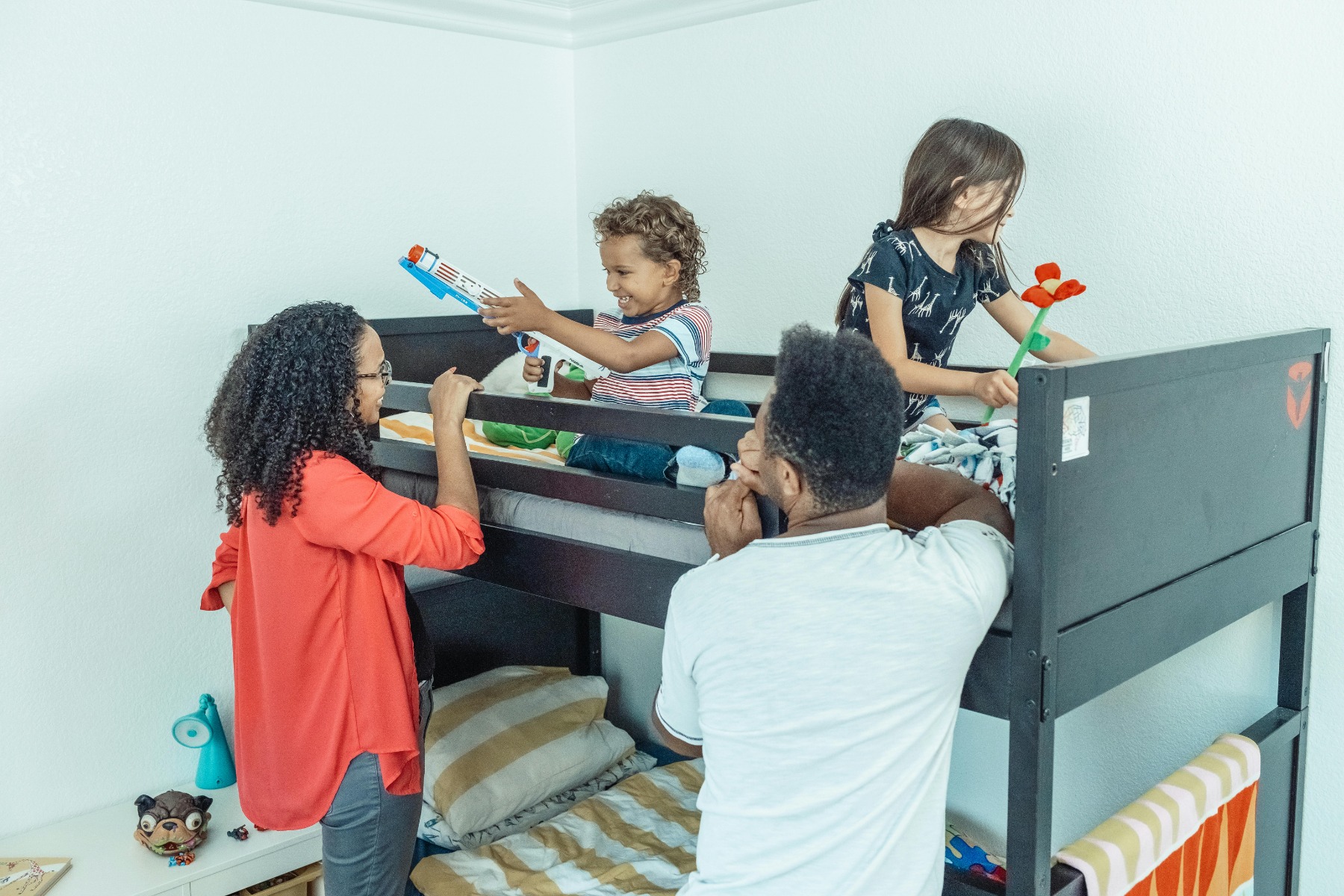 Two adults play with children sitting on a bunk bed in a bright bedroom